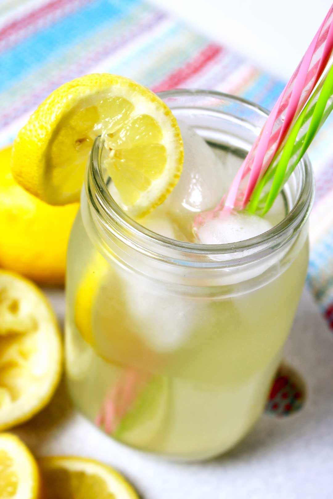 A glass of lemonade in a mason jar with pink and green straws near some lemon slices.