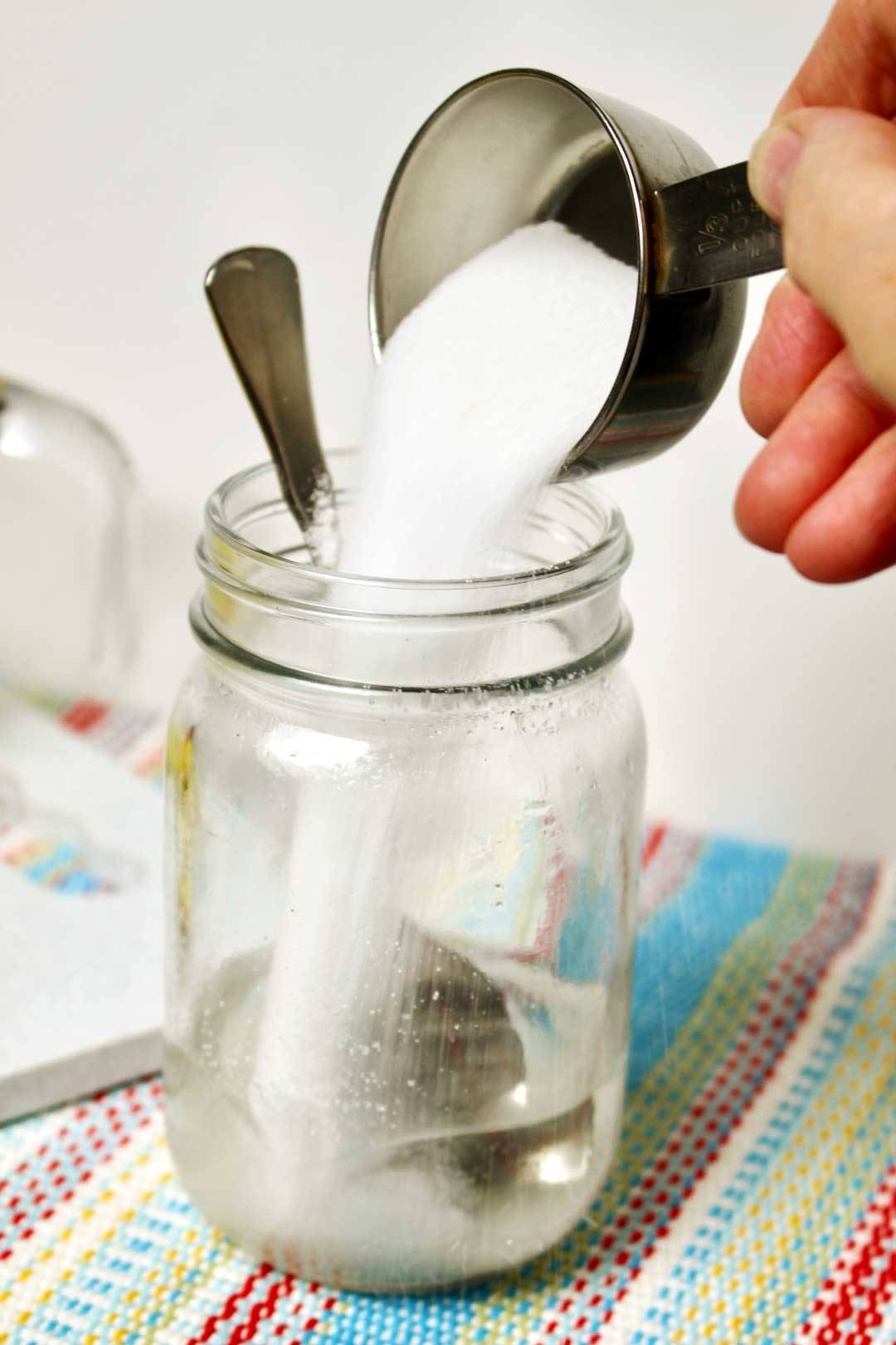 Sugar being poured into water in a mason jar with a spoon near some squeezed lemons.
