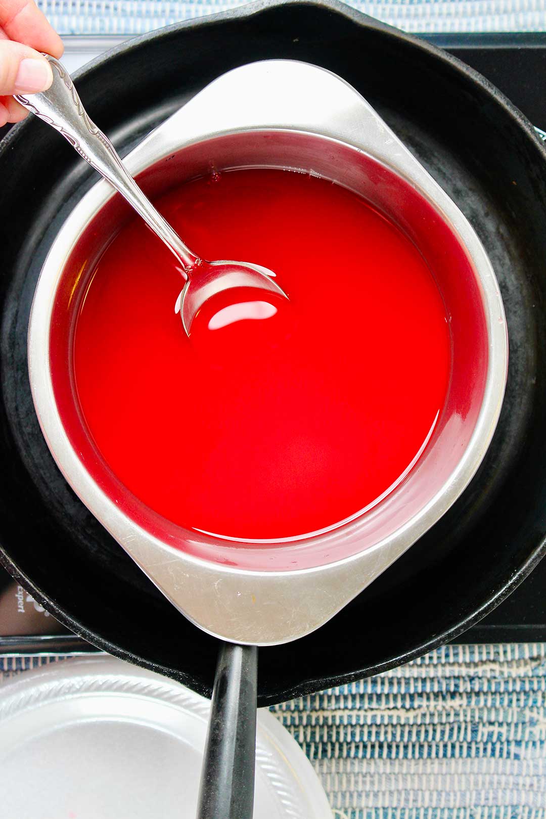 Bowl of melted red wax resting in cast iron pan with person stirring with a spoon.