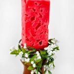 Red rectangular candle on candle holder with greenery and white flowers around it.
