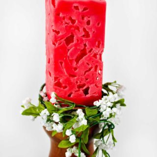 Red rectangular candle on candle holder with greenery and white flowers around it.