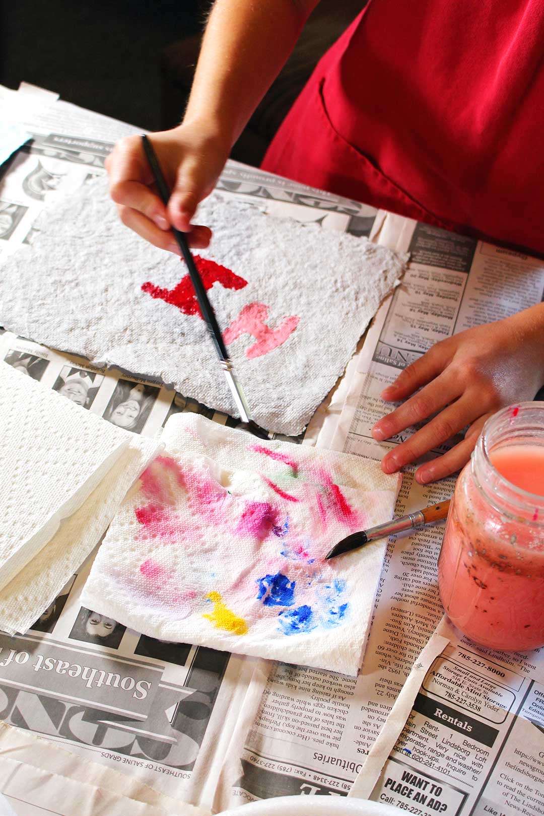 Child in red smock painting on homemade paper, paper towel for blotting and cup for rinsing paintbrushes.
