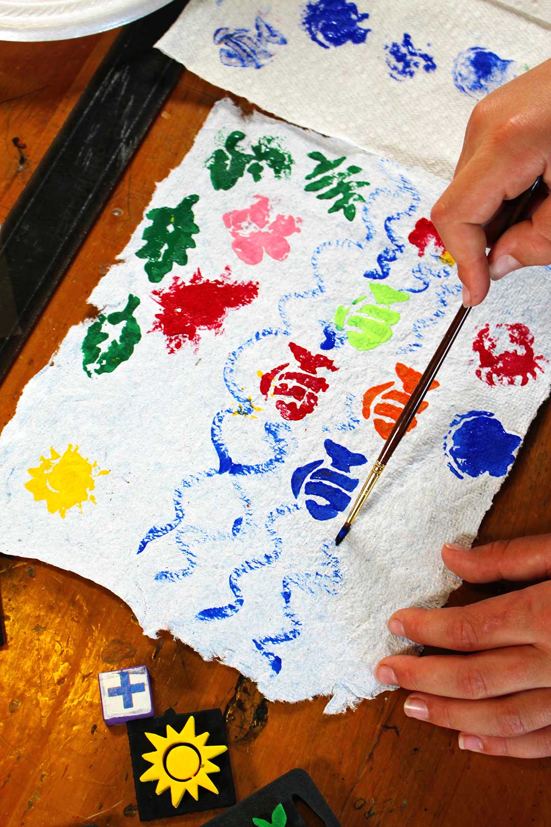 Homemade paper on wooden table. Child's hands painting water on stamped painting of fish.