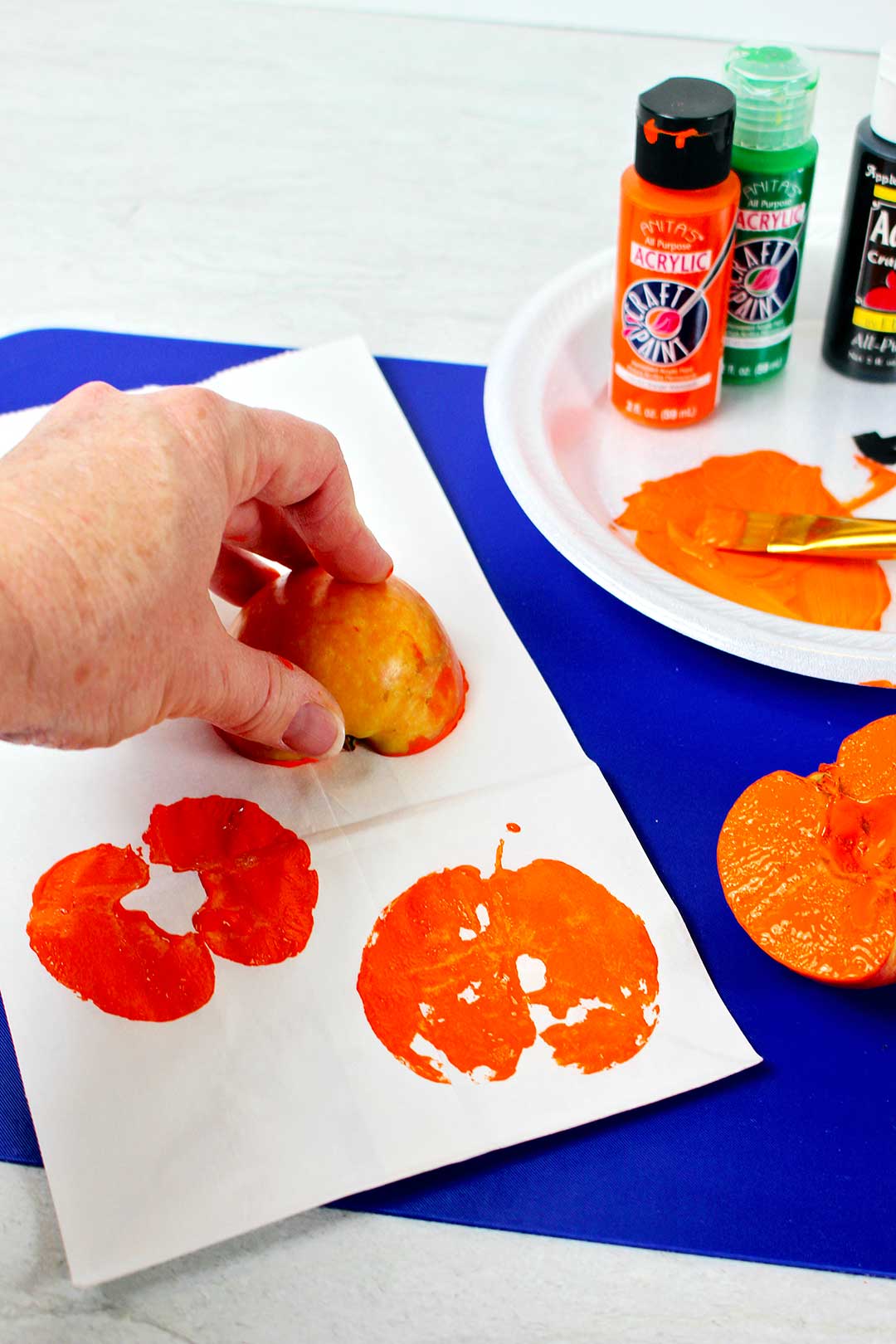 Hand stamping orange pumpkins with an apple half on a white paper bag with supplies in the background.