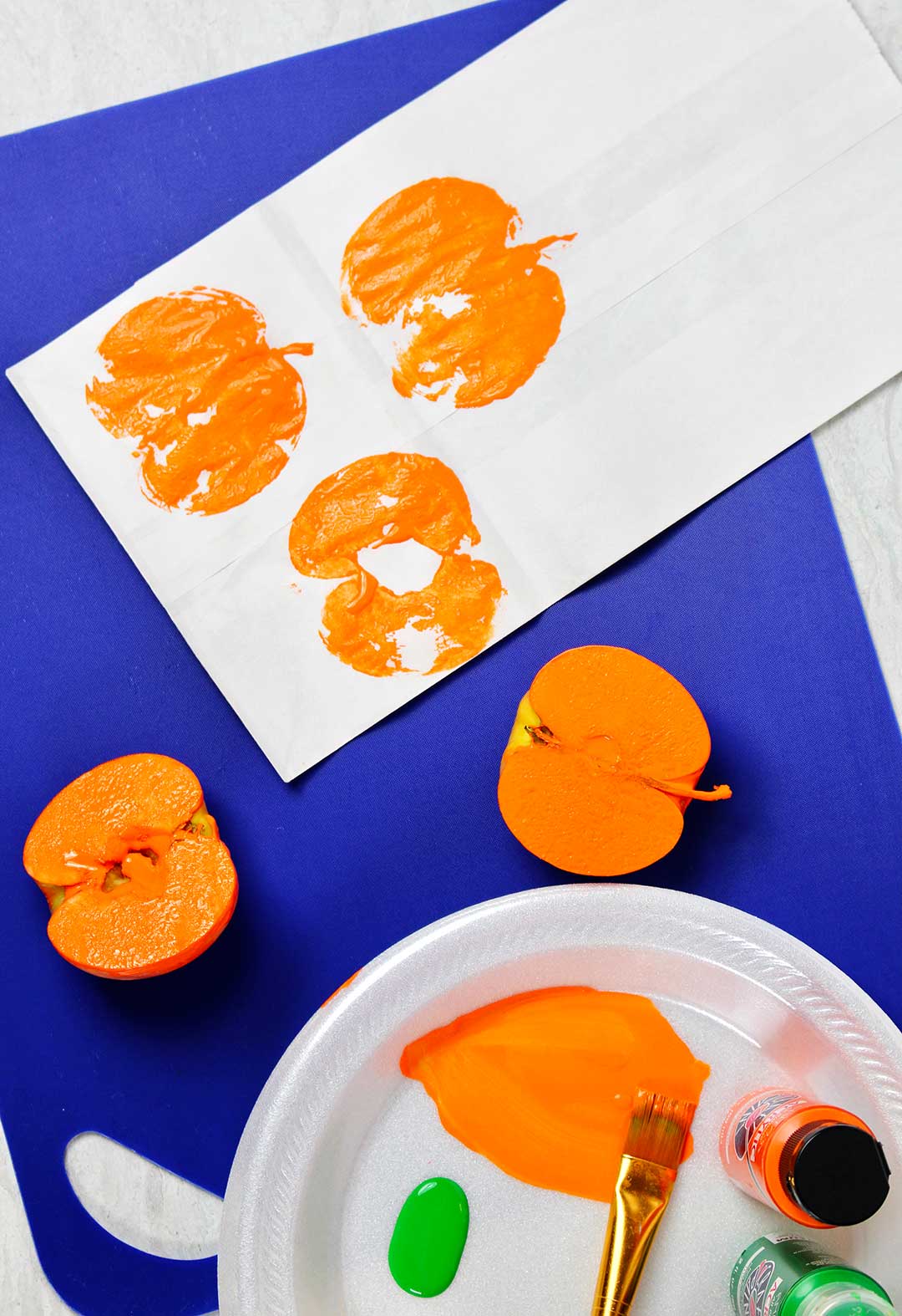 Three pumpkin prints on a white paper bag with halved painted apples and orange and green paint.