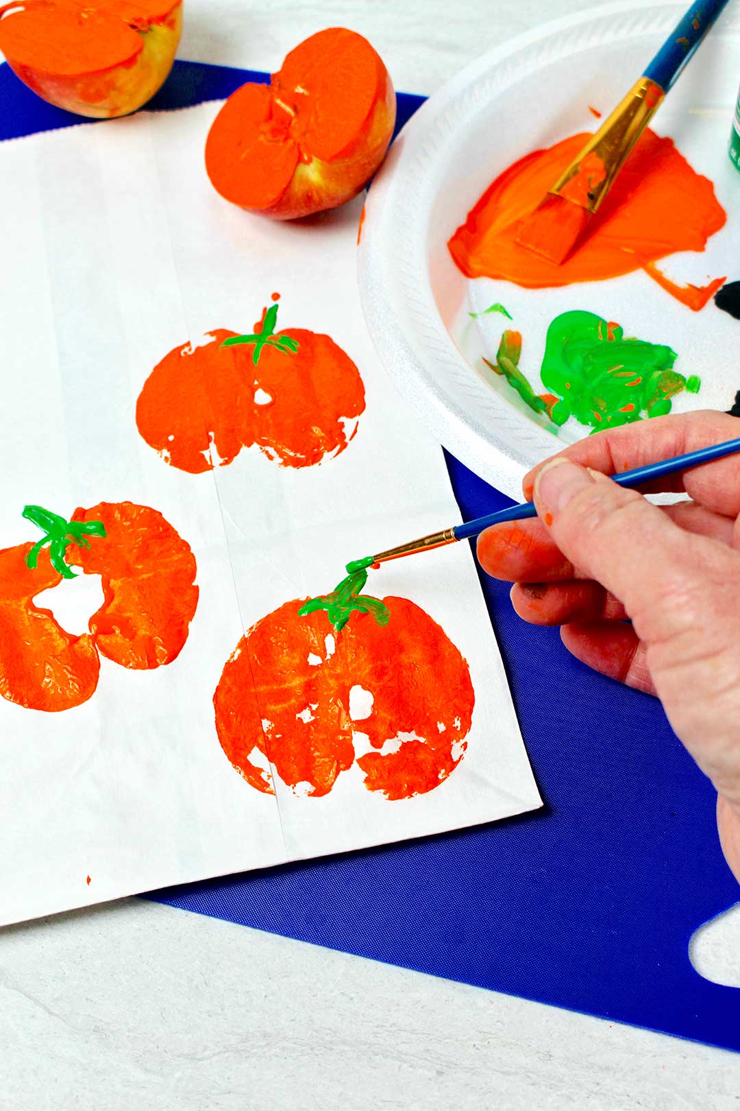 Person painting green stems on orange pumpkin prints with painted apple halves and paint nearby.