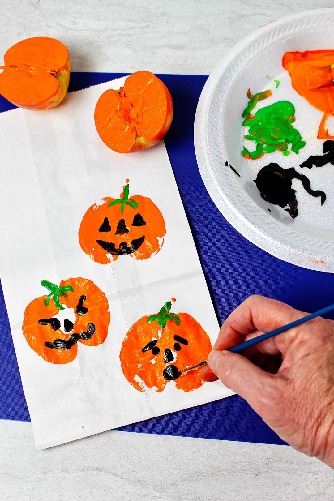 Person painting black jack-o-lantern faces on pumpkin prints on white paper bag.