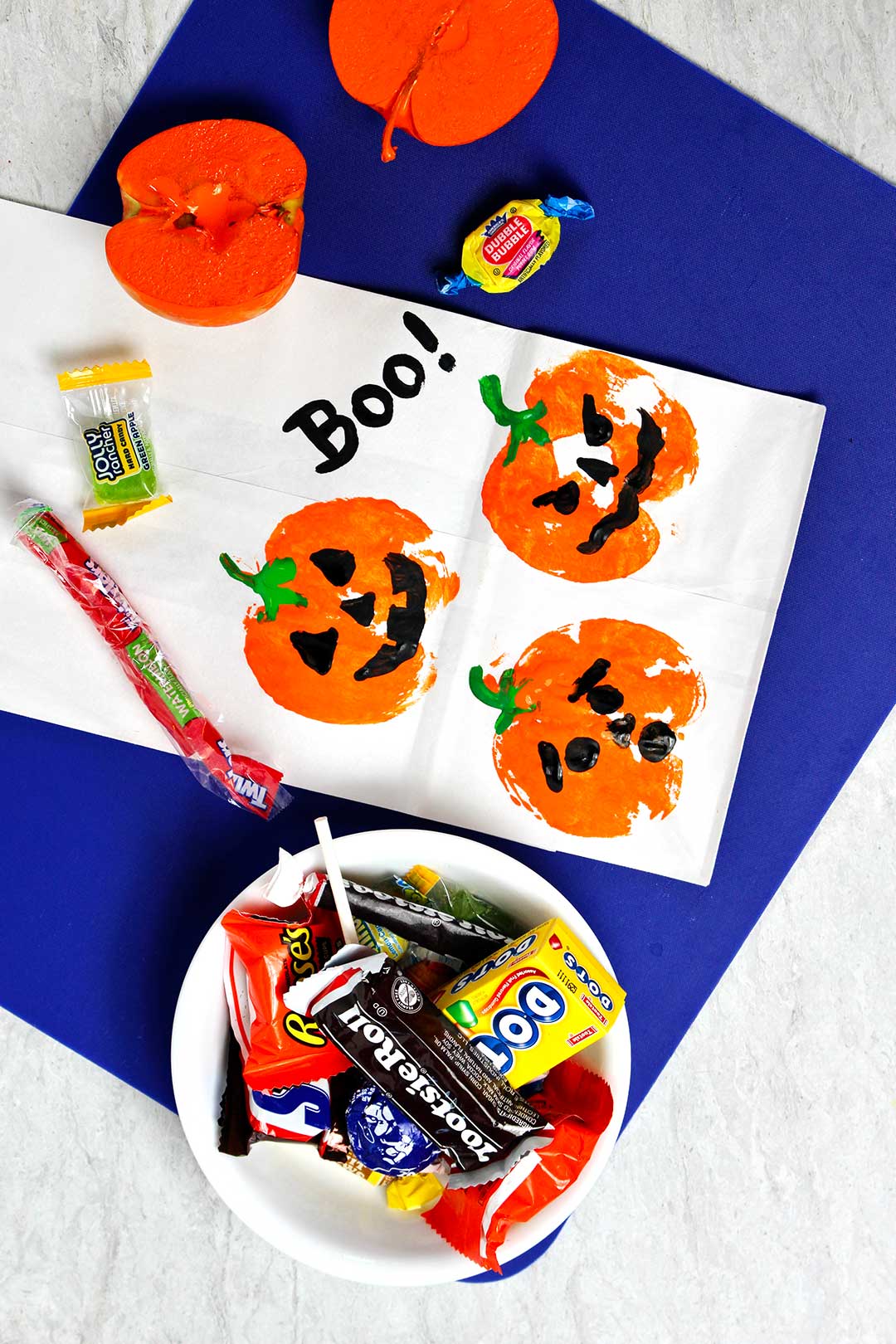 White paper bag with pumpkin prints on top of blue cutting board next to a bowl of Halloween candy.