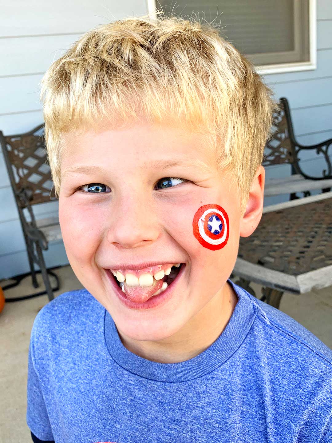Boy with blonde hair with Captain America shield face painted on cheek making silly face.