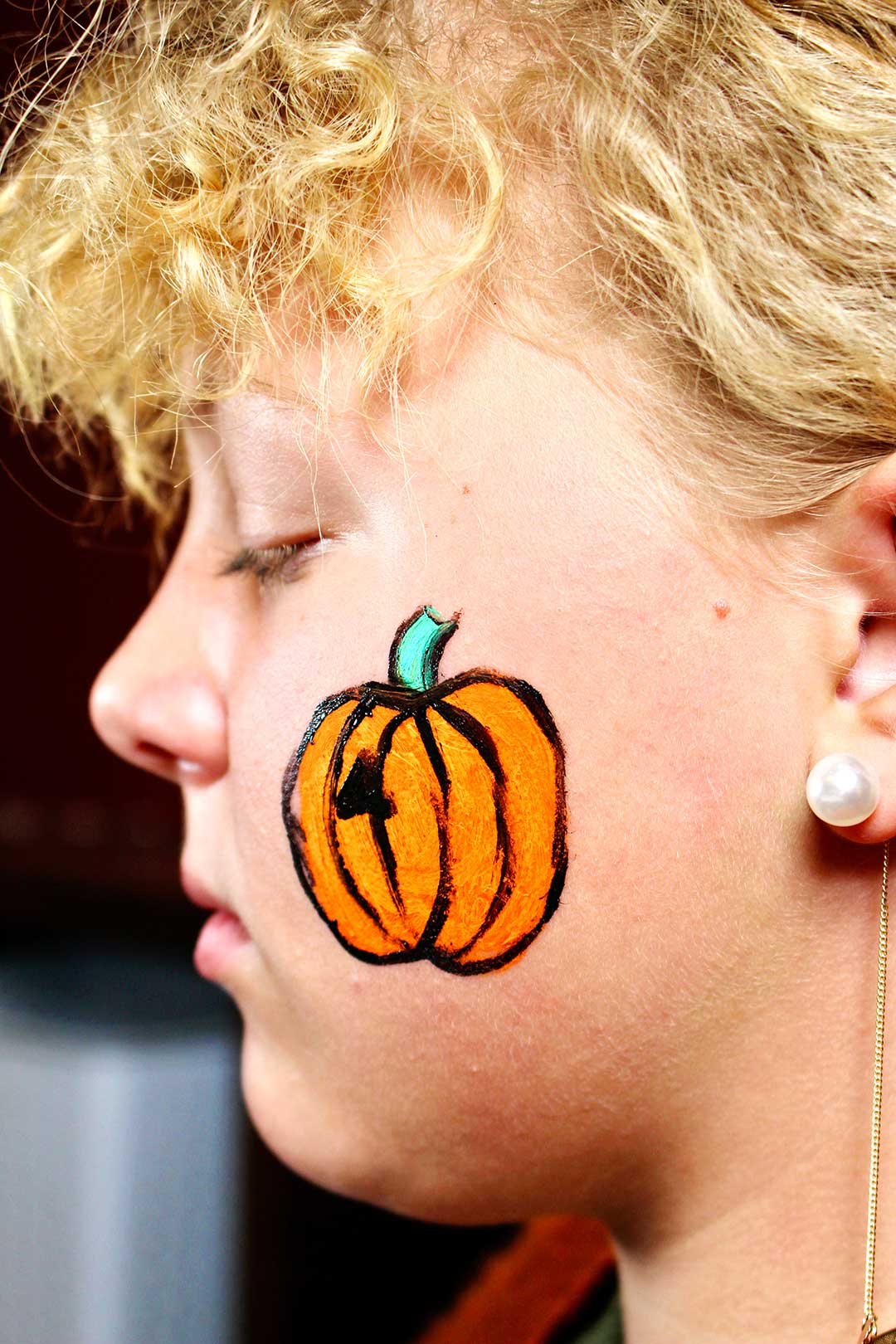 Profile of girls face with more detailed painting of orange pumpkin with green stem and black outline.