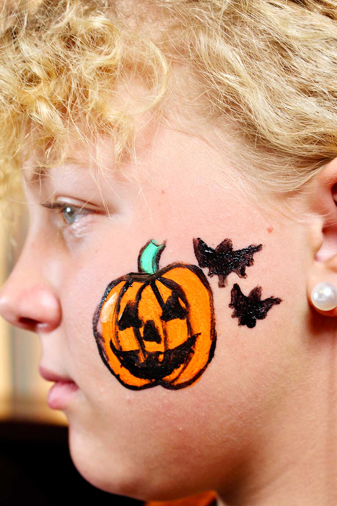 Profile of girl with finished jack-o-lantern and two black bats on cheek.