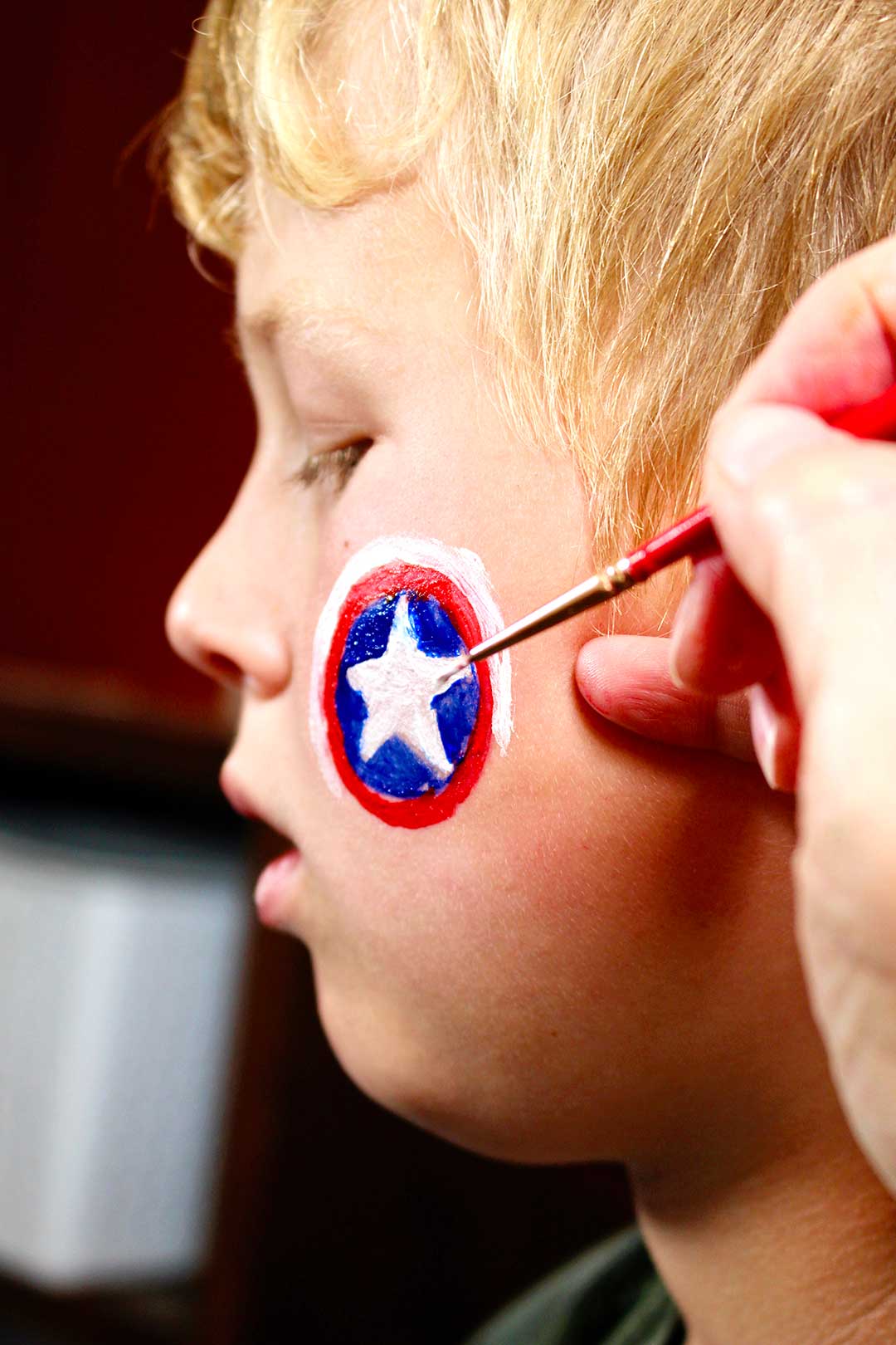 Profile of boy with hand painting details of Captain America shield on cheek.