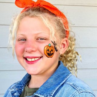 Girl with blonde curly hair smiling at camera with jack-o-lantern face painting on cheek.