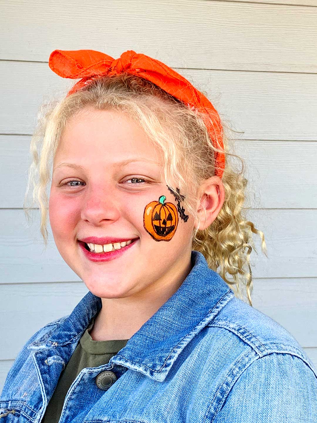 Girl with blonde curly hair smiling at camera with jack-o-lantern face painting on cheek.