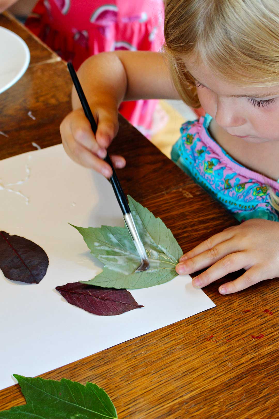 Girl with blonde hair painting the back of a leaf with paste for her leaf collage.