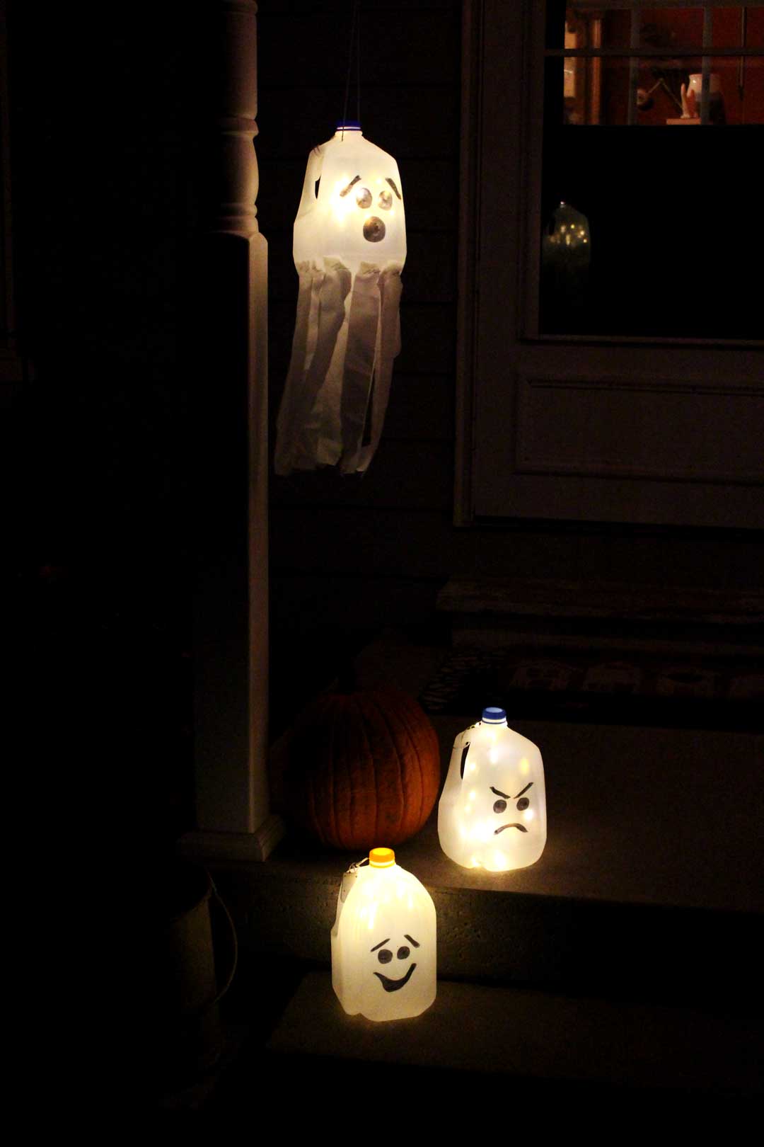 Recycled milk jugs with faces drawn on them to create glowing ghost luminaries hanging on a porch at night.