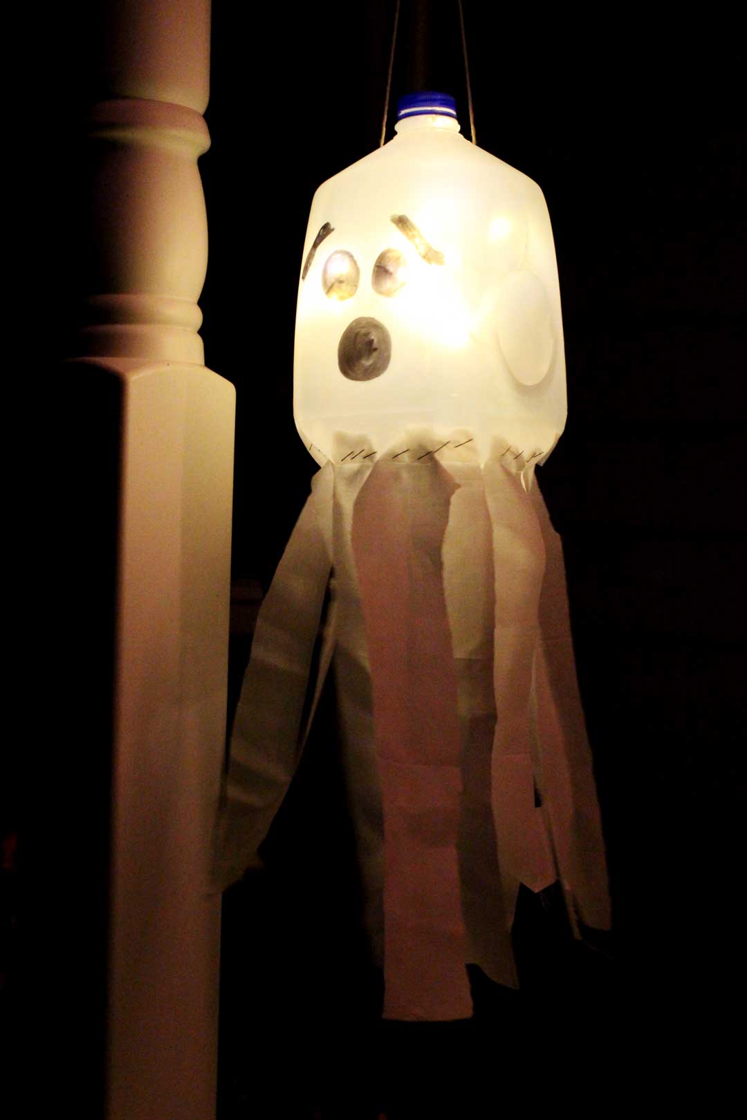 A recycled milk jug with a face drawn on it to create a glowing ghost luminary hanging on a porch at night.