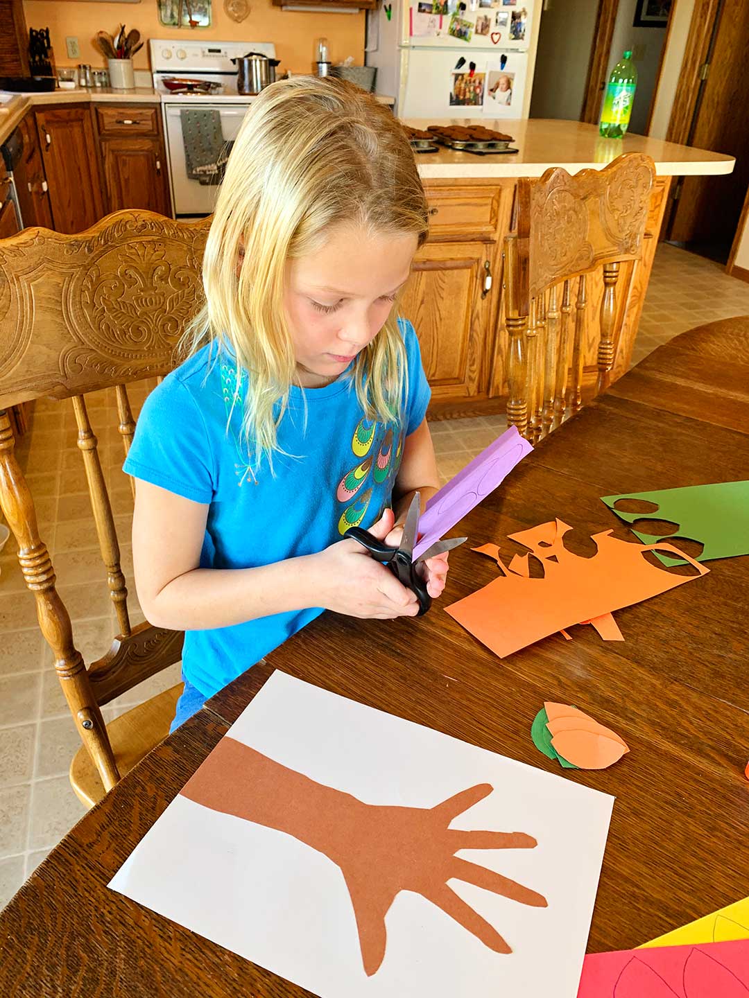 Young girl wearing blue shirt sitting at kitchen table cutting out leaves out of colored construction paper.