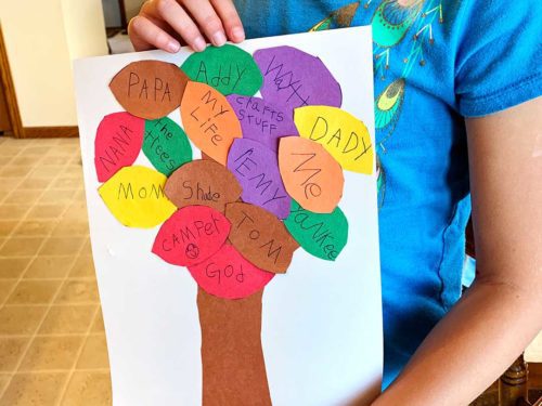 Girl with blonde hair wearing a blue t-shirt holding completed Blessing Tree craft complete with colored leaves with names written on them.