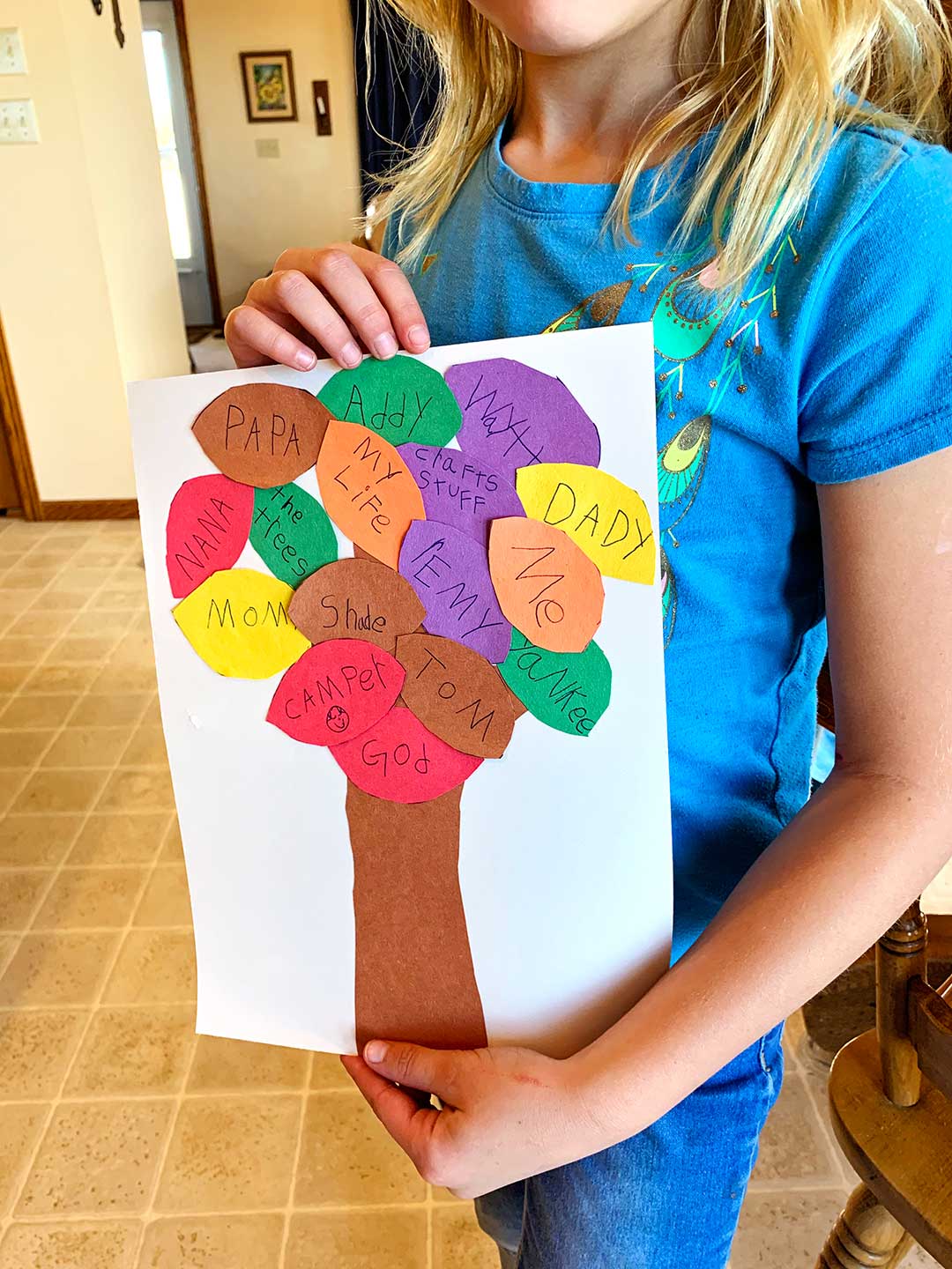 Girl with blonde hair wearing a blue t-shirt holding completed Blessing Tree craft complete with colored leaves with names written on them.