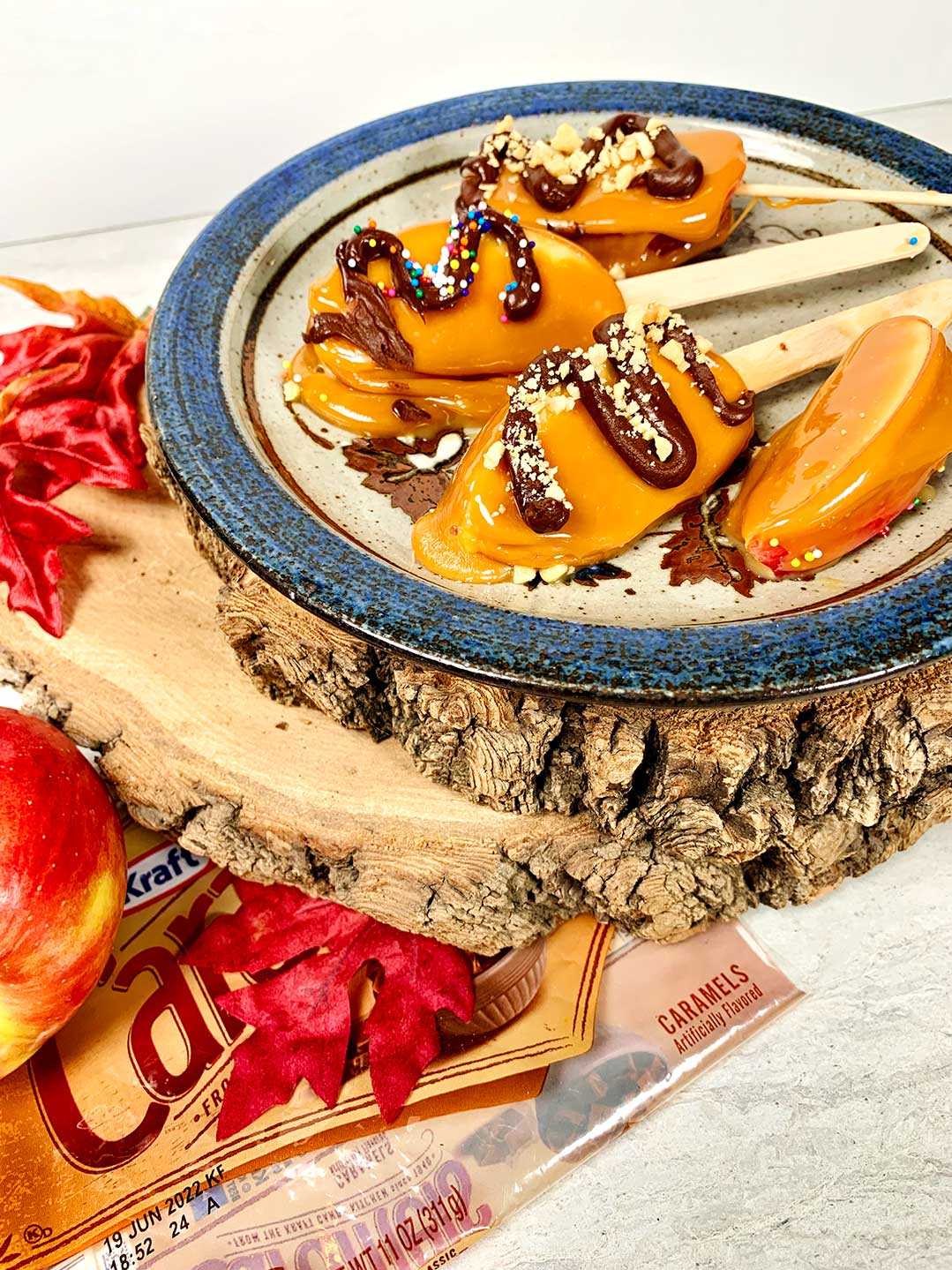 Plate of decorated caramel apple slices on a stick on wood slices with leaves and apples nearby.