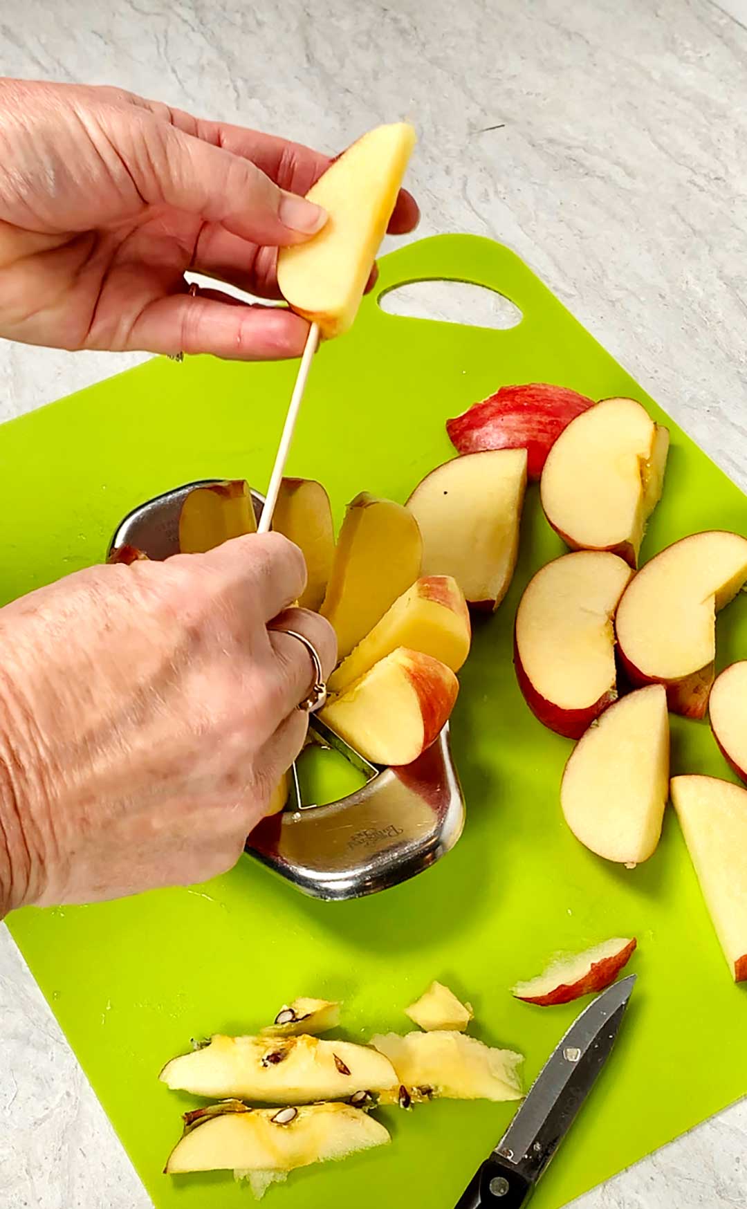 Person sticking stick through apple slice with other apple slices nearby on bright green cutting board.