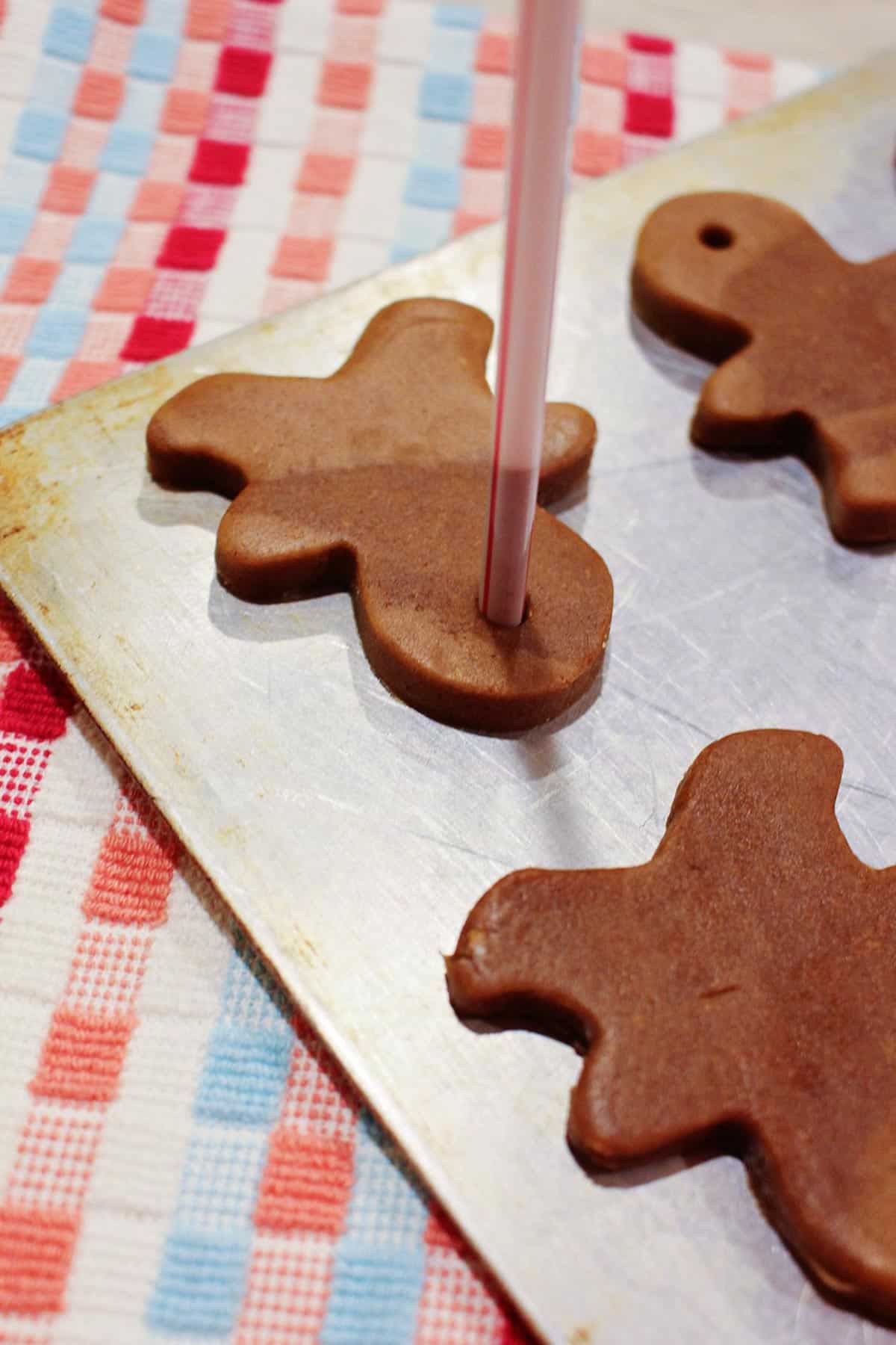 A straw poking a hole in the top of an unbaked gingerbread cookie on a baking sheet.