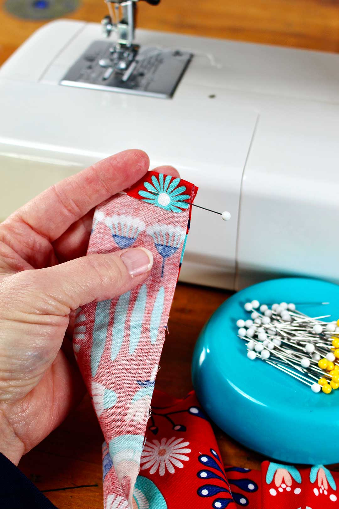 Straight pin holding edge of a strip of fabric for sewing. Sewing machine and straight pins nearby as person shows how to make a scrunchie.