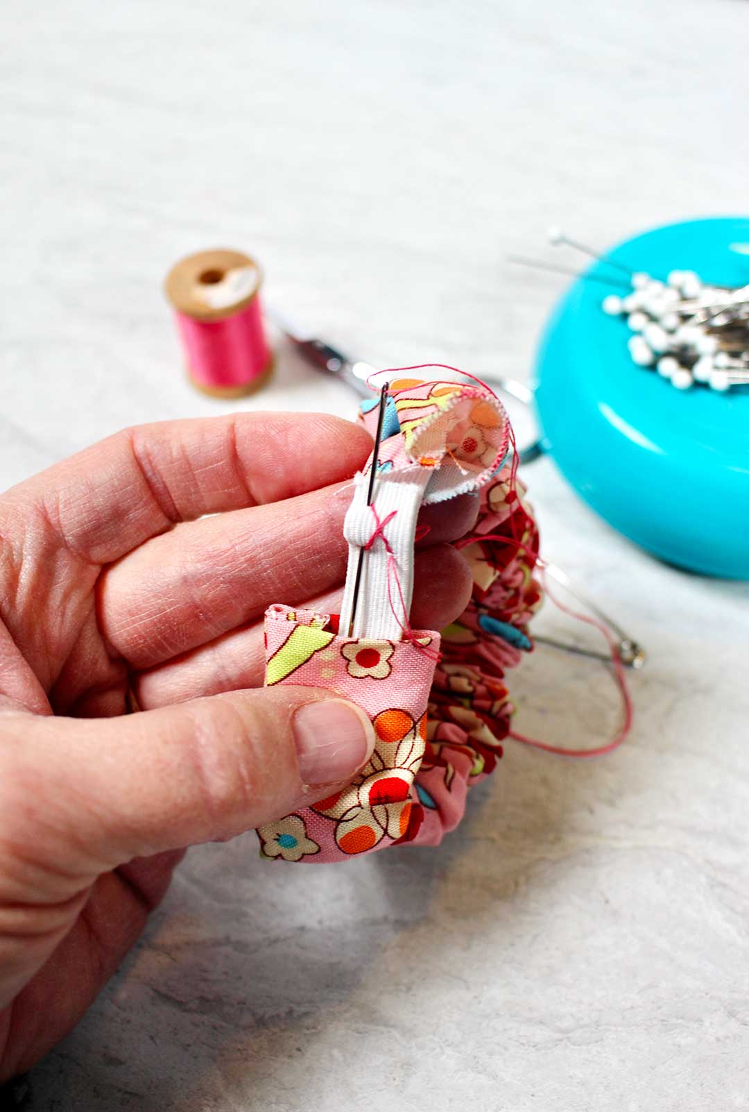 Person holding scrunchie showing how to secure the fabric to the elastic of the scrunchie. Pins and pink thread nearby.
