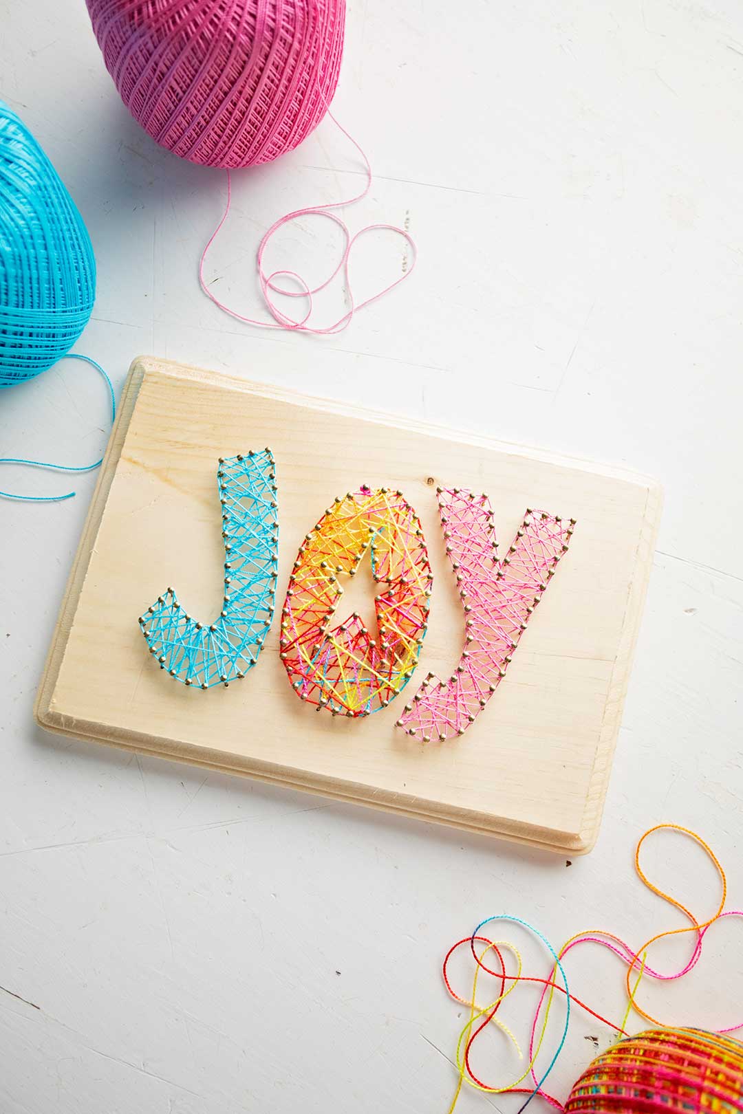 Finished "joy" string art on light wood with blue, pink and multi colored string near by.