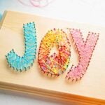 Colorful string woven around nails on a wooden board to spell the word, "Joy".