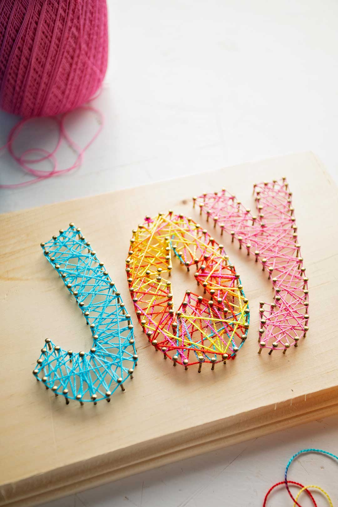 Angle close up of finished "joy" string art in blue, multi color and pink string.