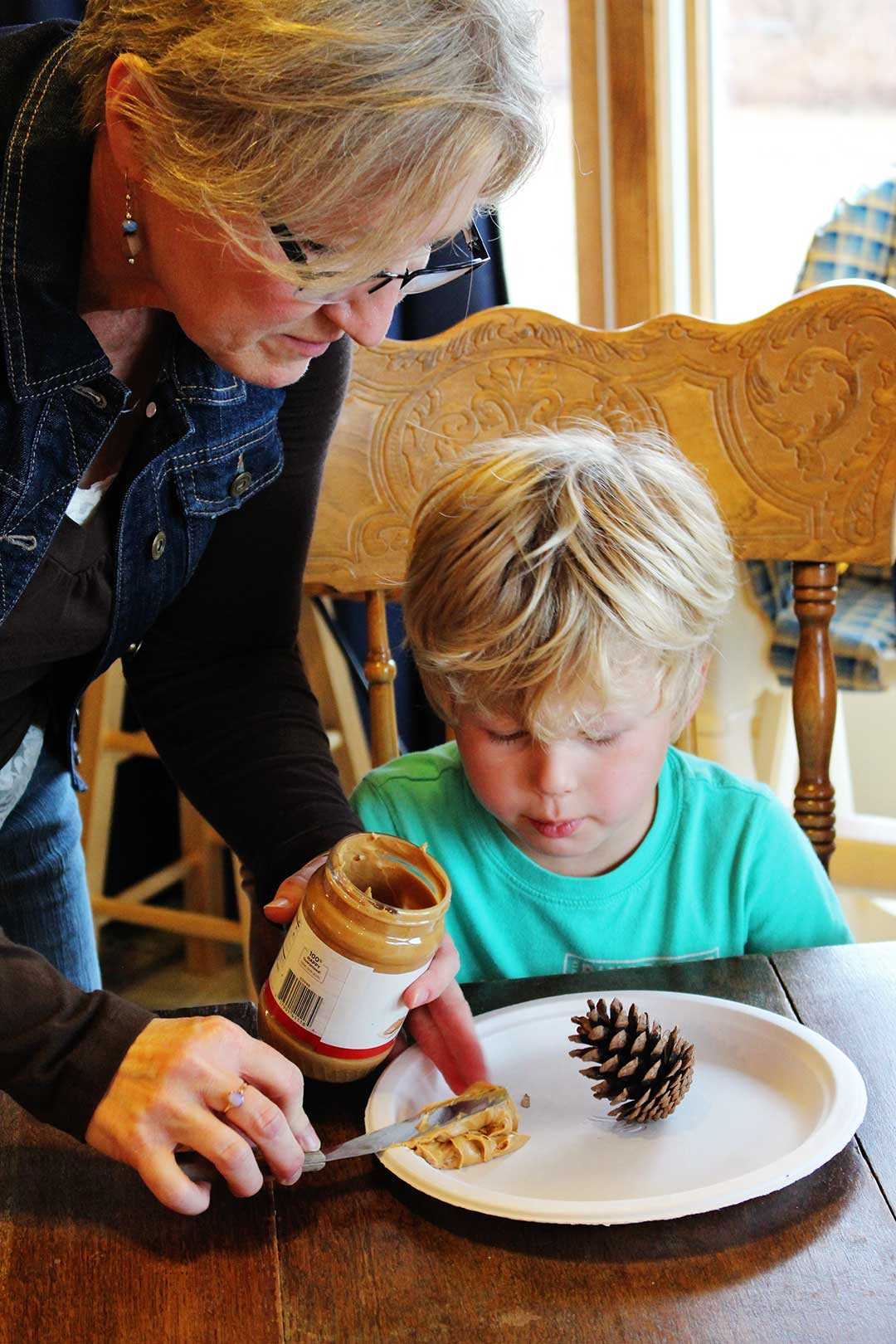 Blonde woman helps small boy by adding a dollop of peanut butter on to a paper plate for his bird feeder project.