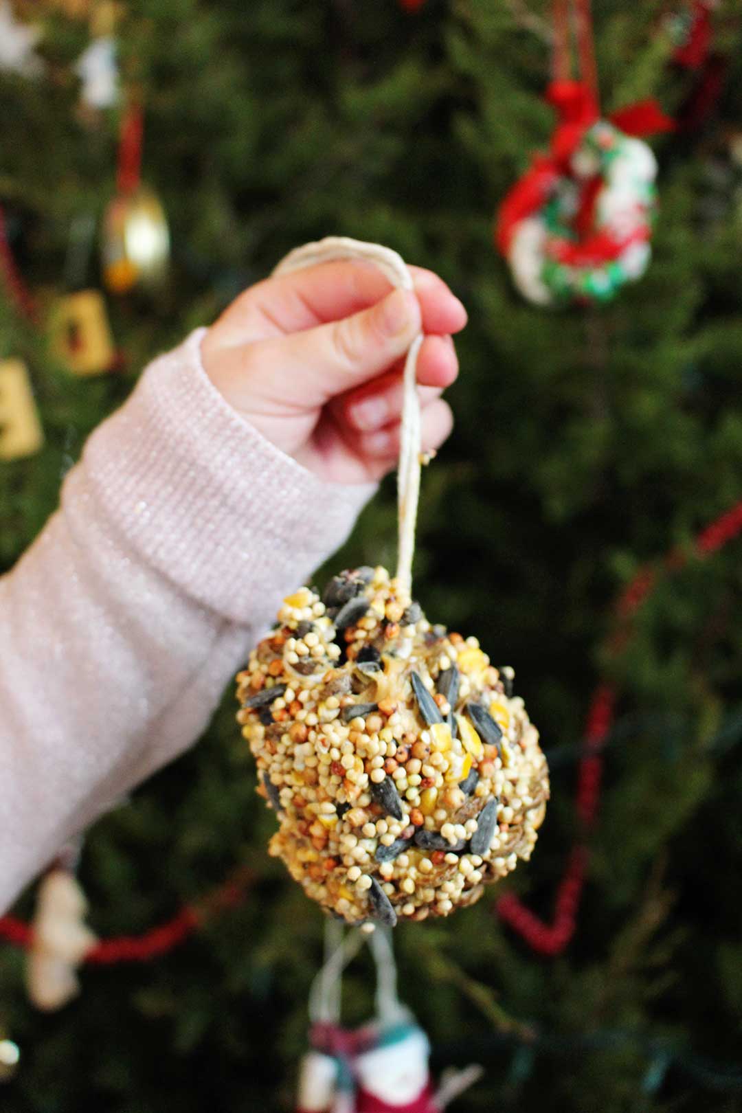 Little hand holding finished pine cone bird feeder in front of Christmas tree