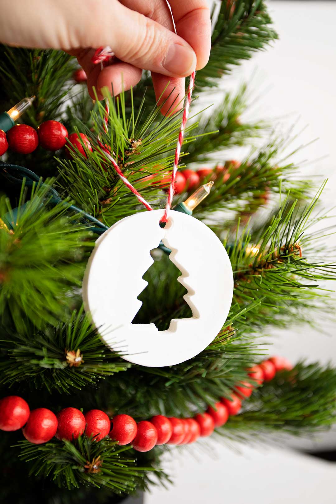Person hanging circular cornstarch ornament with tree cut-out on a Christmas tree with white lights and red wooden garland.