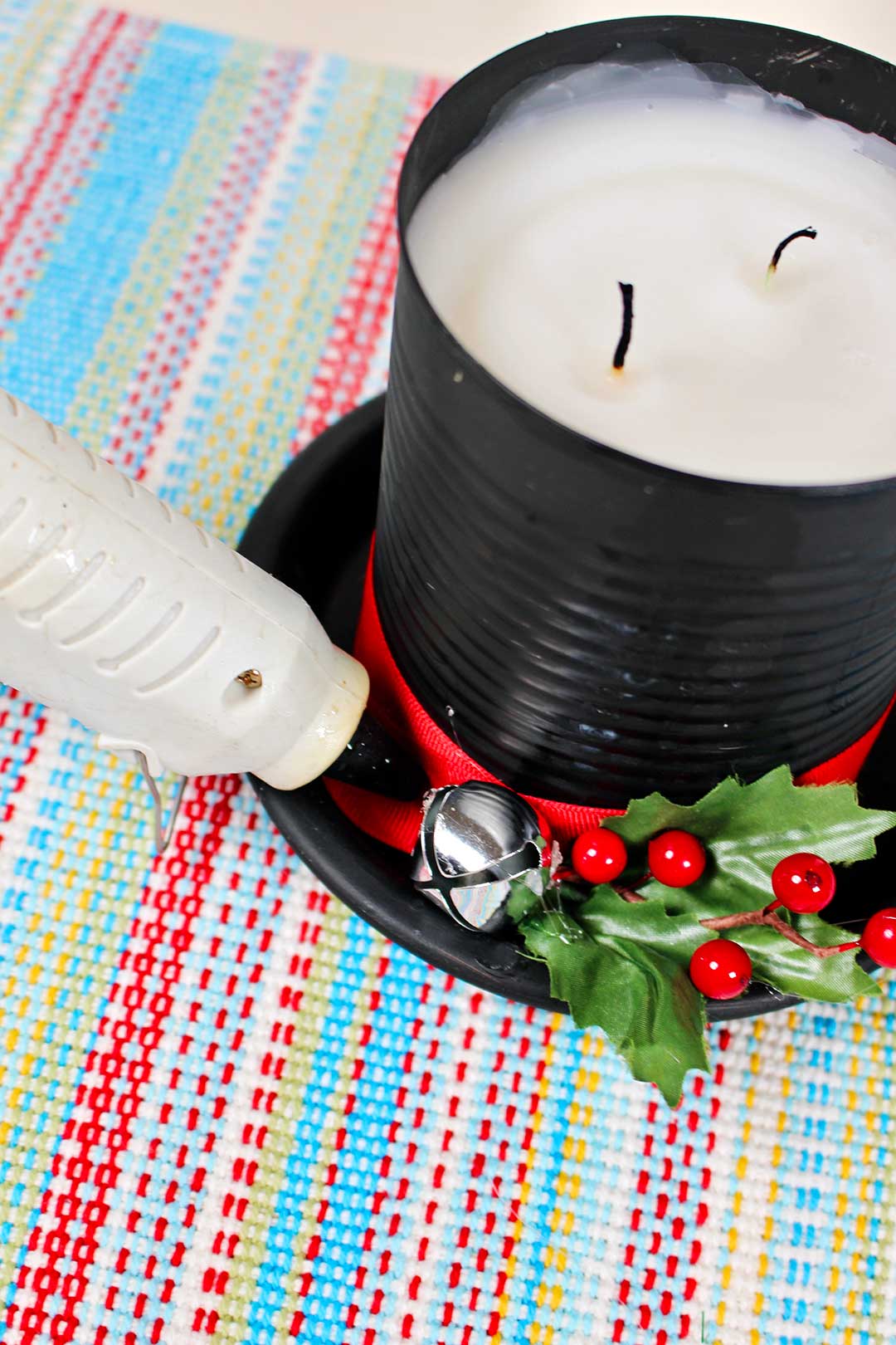 DIY Frosty The Snowman Candle resting on multi colored placemat with person glueing a bell with glue gun.