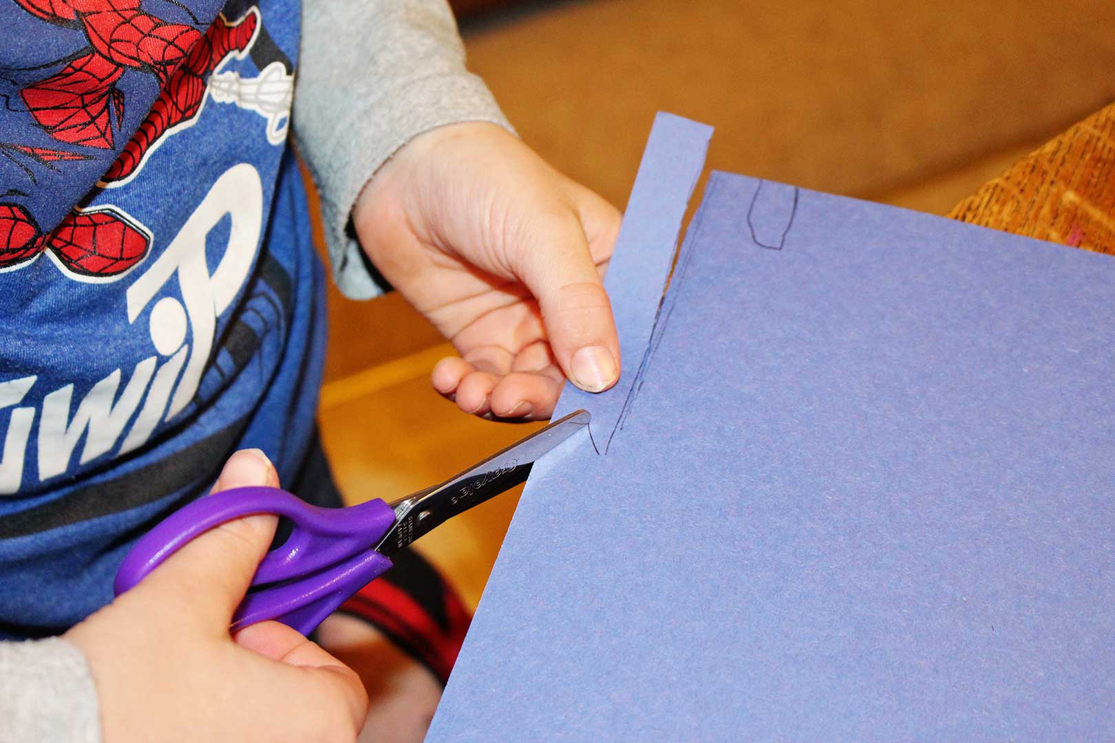 Child in a Spiderman shirt cutting blue construction paper to be used a a scarf for the 3-D paper plate snowman craft.