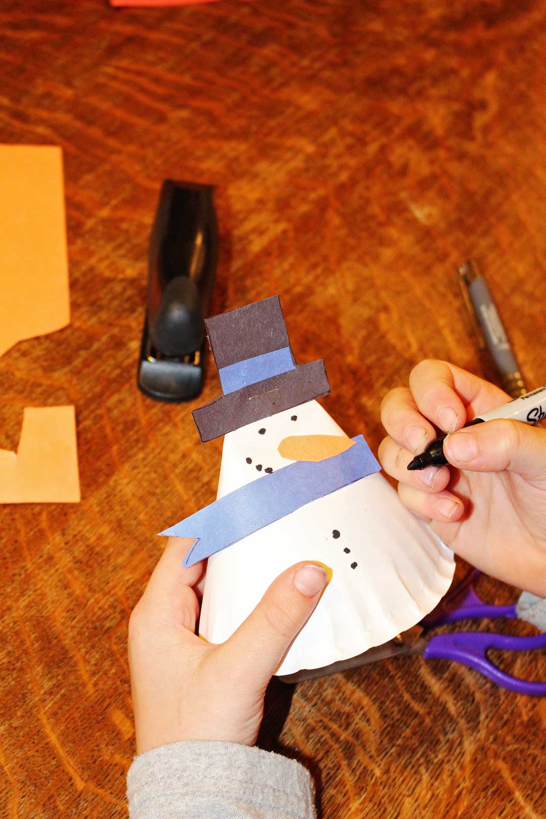 Person drawing black buttons on 3-D paper plate snowman with a Sharpie marker with various supplies near them on a wooden table.