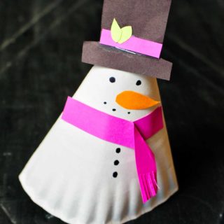 Finished 3-D paper plate snowman craft sitting on a black marble surface. Snowman has fuchsia scarf and decoration on its black top hat.