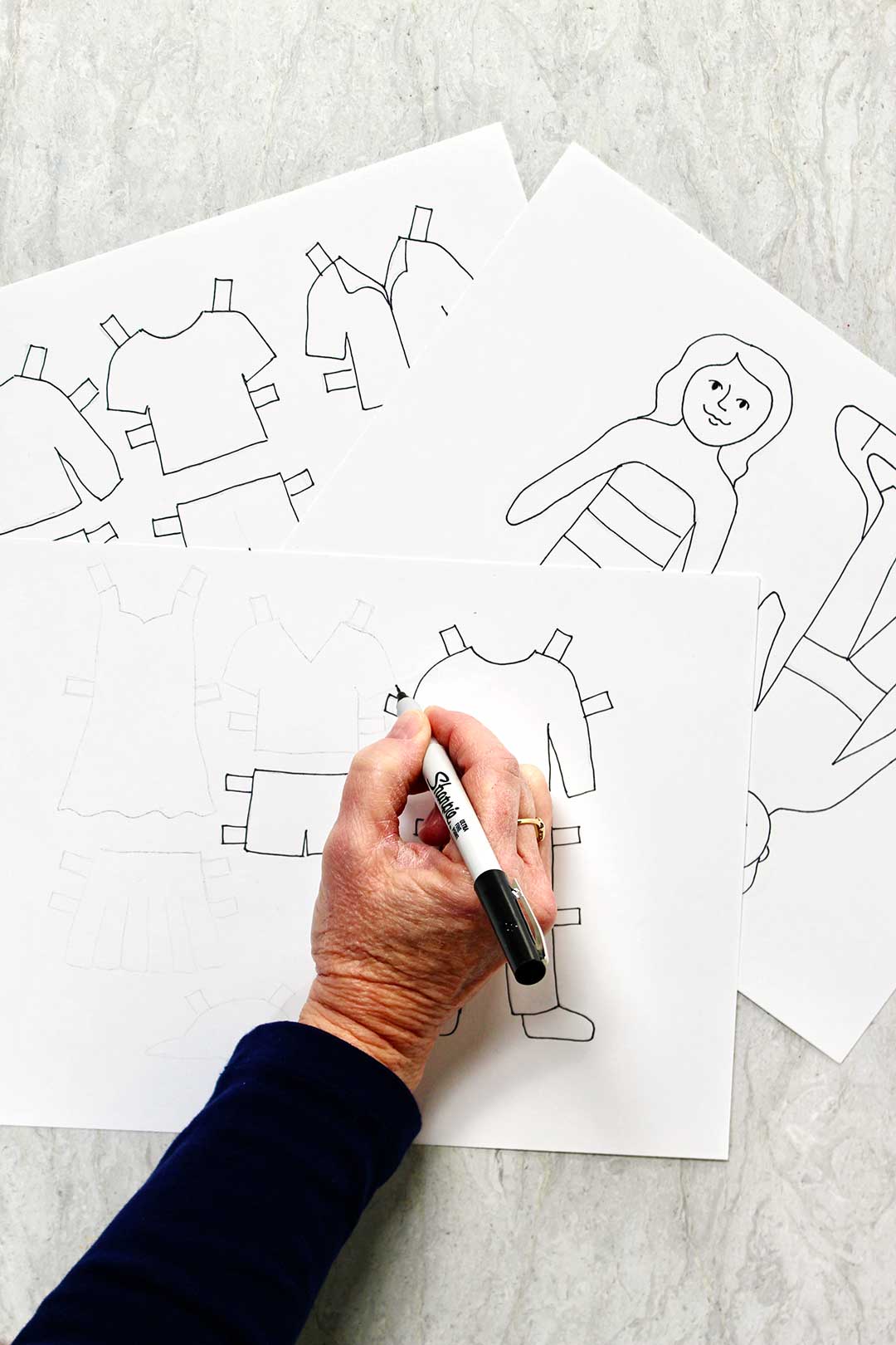 Person drawing outfits for paper dolls on white paper with a black Sharpie.