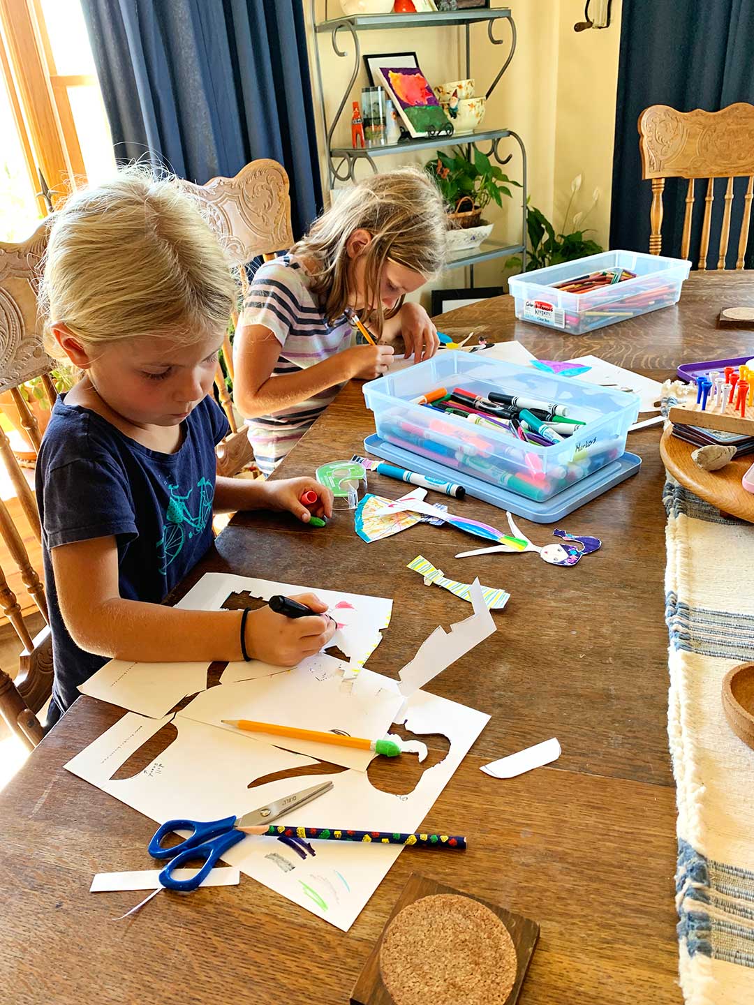 Two girls at a kitchen table coloring and cutting their own paper dolls with markers and colored pencils.