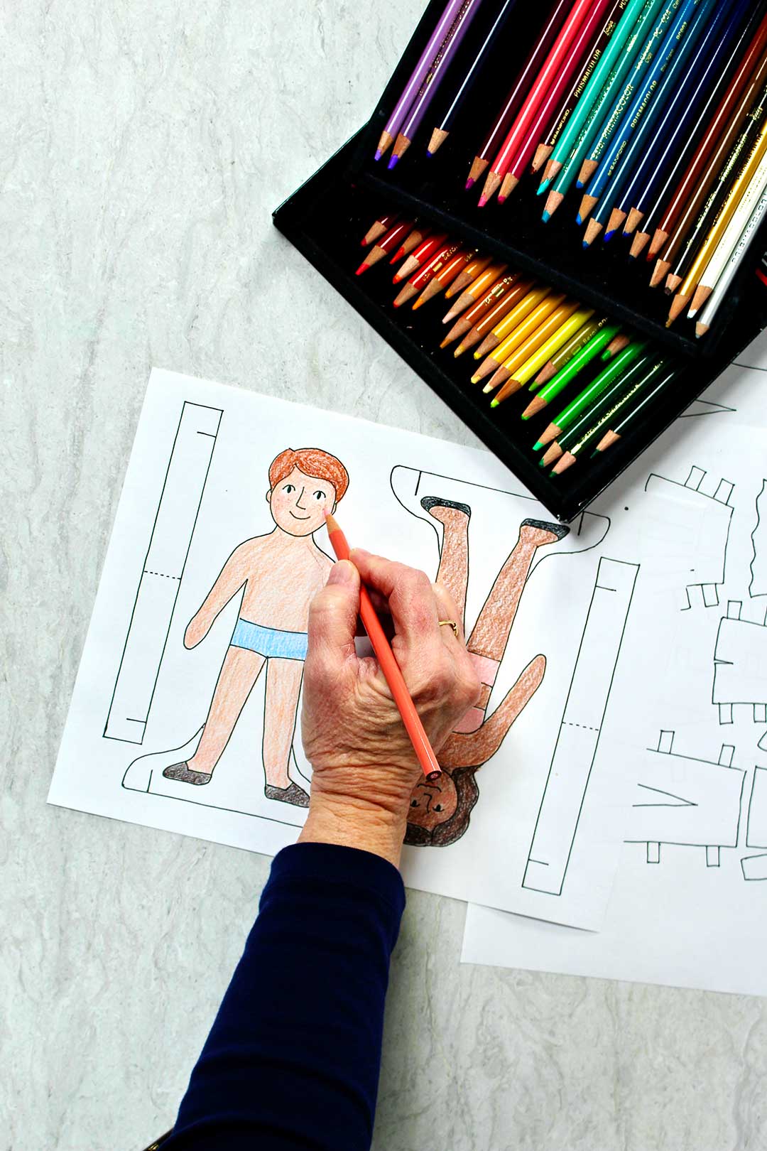 Person coloring paper dolls with orange colored pencils. A rainbow of colored pencils nearby.