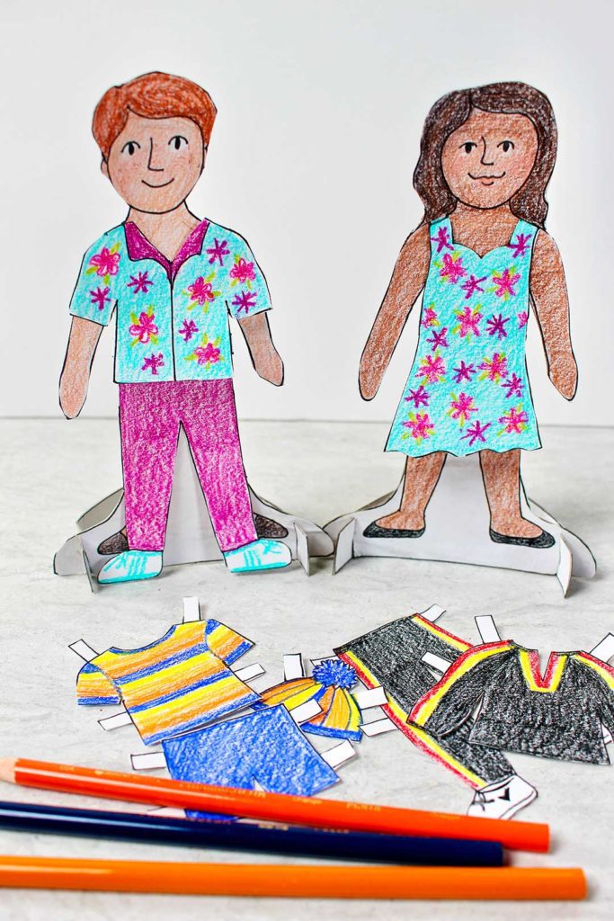 Poupées en papier homme et femme, colorées et habillées sur leurs socles.  Crayons de couleur et autres vêtements de couleur à proximité.