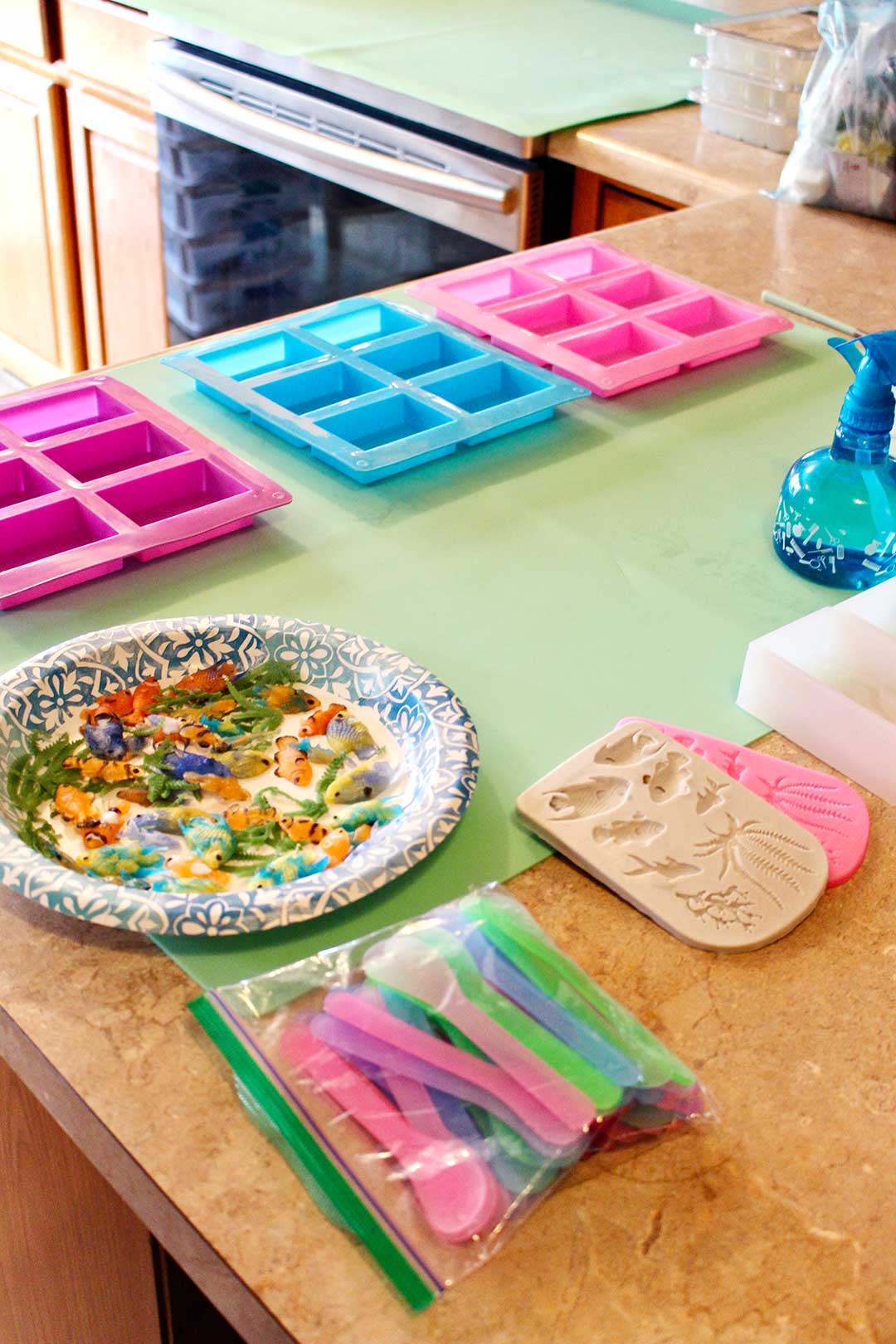 Soap making supplies on a counter. Rectangular soap molds, colorful plastic spoons and a paper plate with plastic tropical fish.