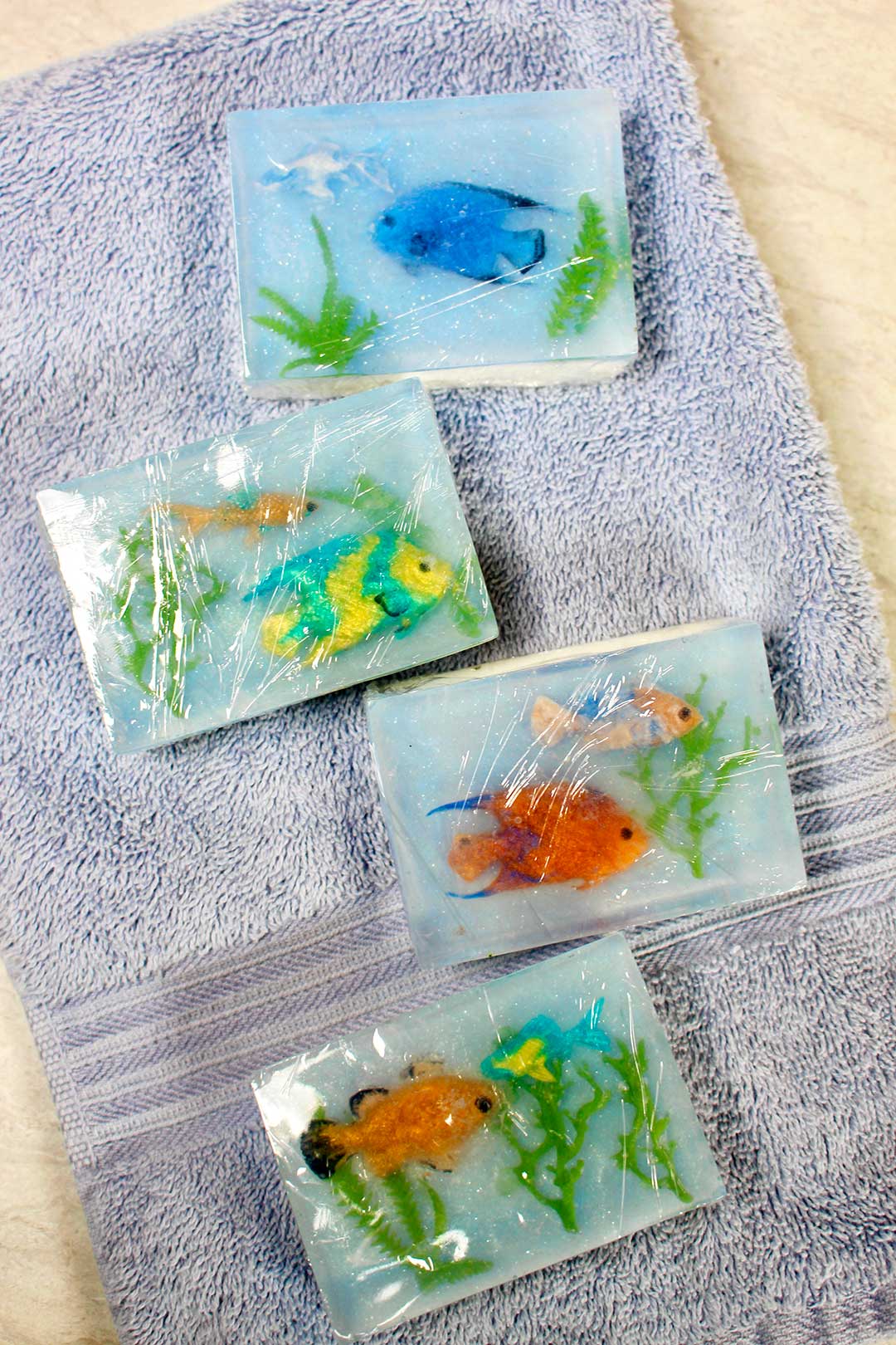 Four completed soaps, packaged in plastic wrap resting on a blue bath towel.