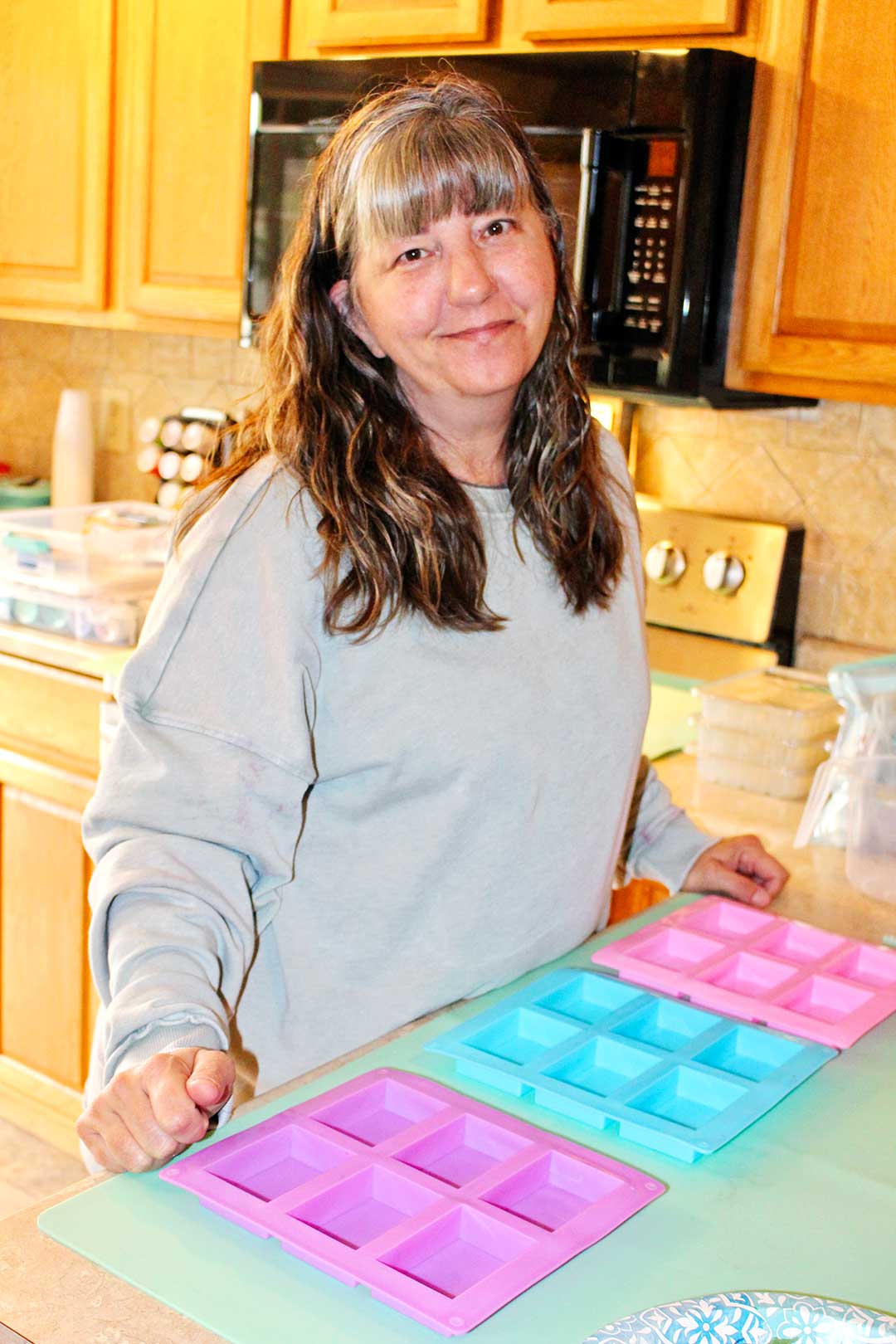 Woman smiles at camera standing at counter with three colorful rectangular soap molds.
