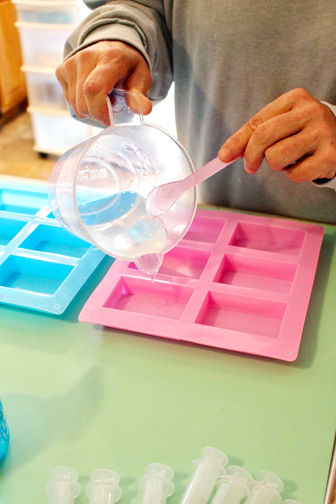 Hands pouring melted soap from measuring cup into pink silicone rectangular soap mold.