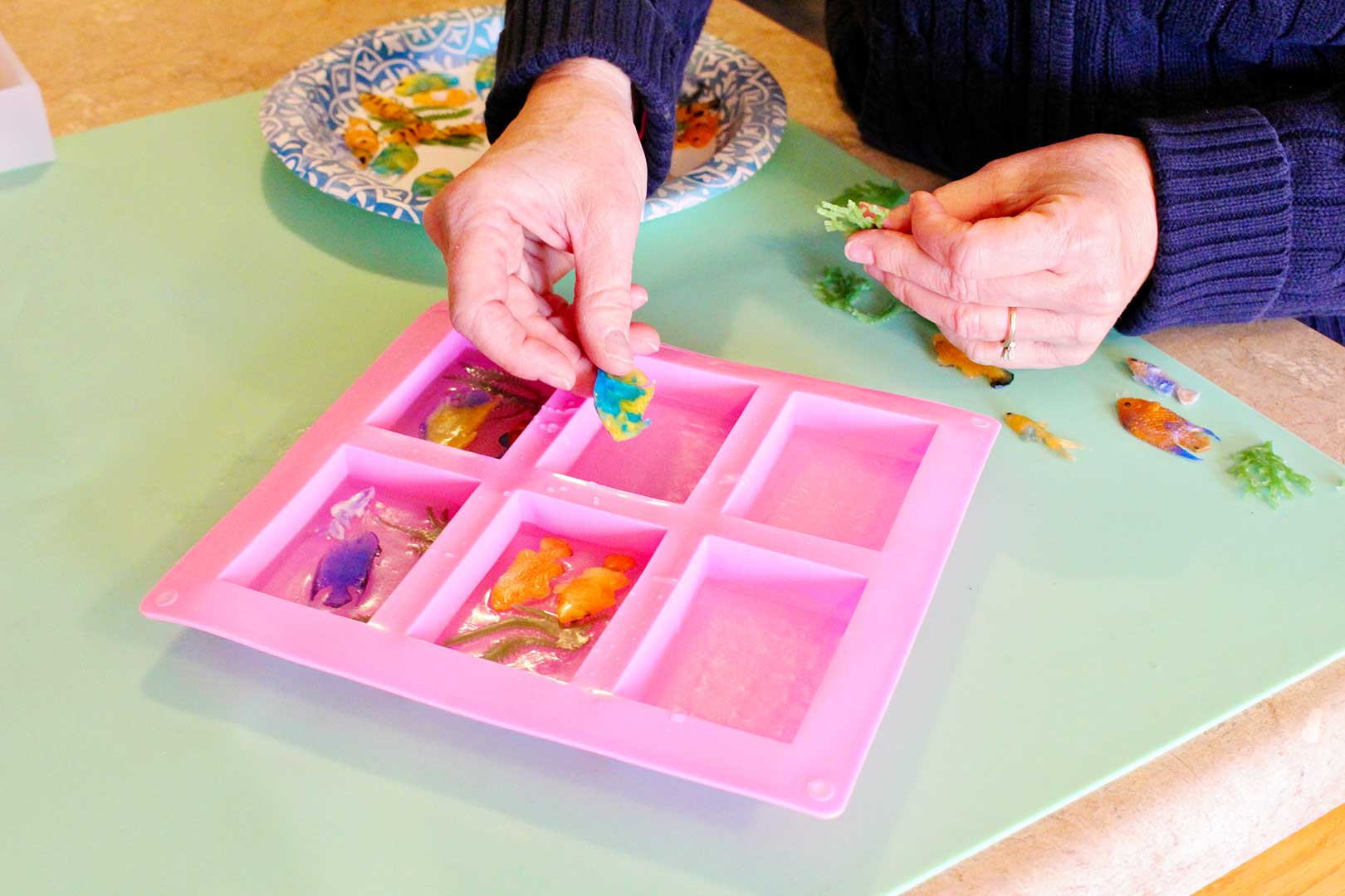 Hands placing plastic tropical fish into clear soap layer inside the pink silicone rectangular soap molds.