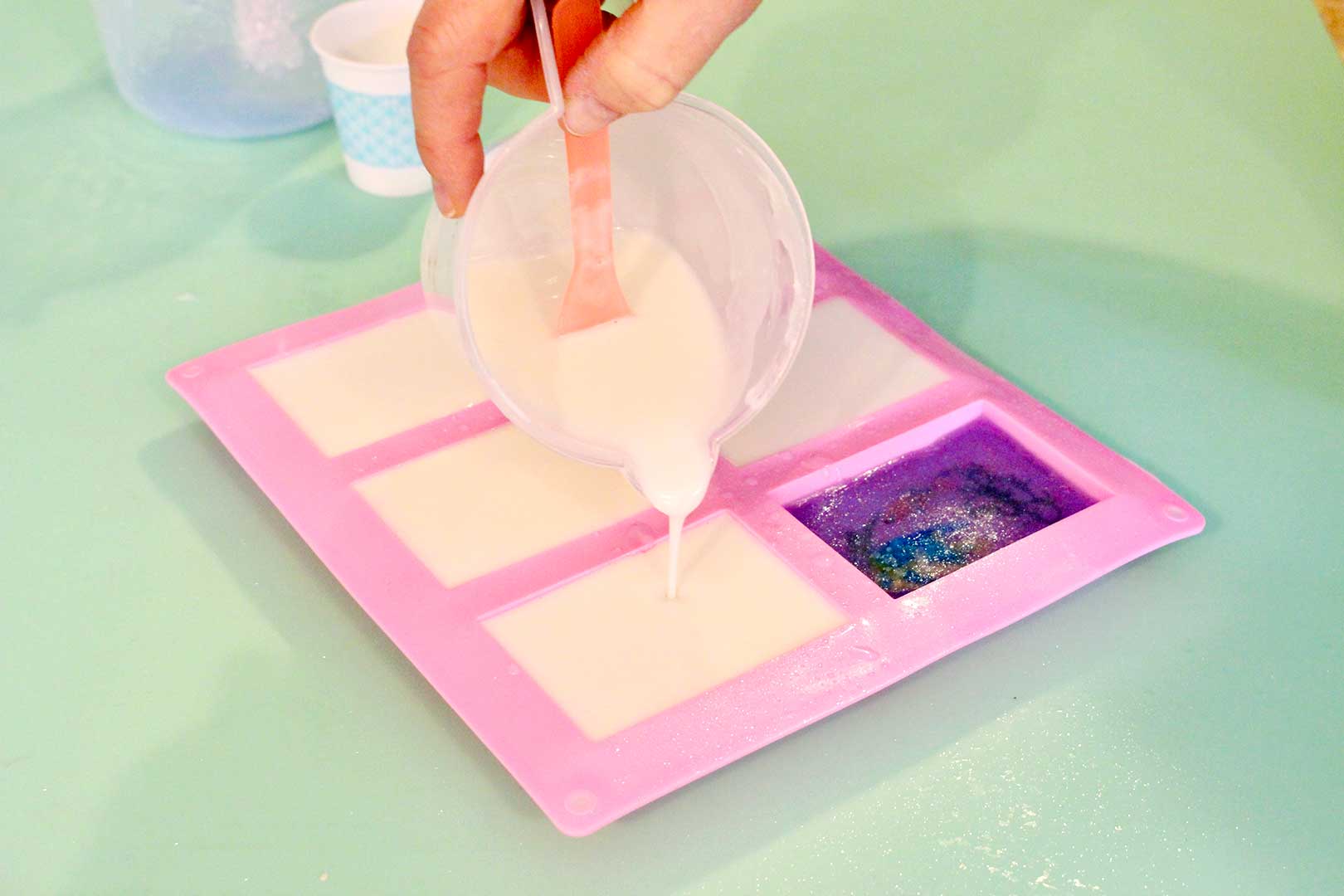 Hand pouring off-white colored melted soap from a measuring cup onto clear fish layer of soap resting on a mint green background.