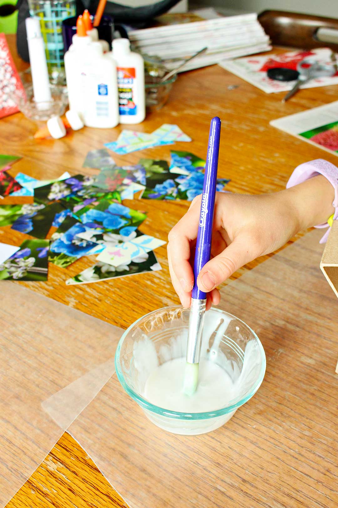 Child dipping paintbrush into a glass bowl full of Mod Podge with other decoupage supplies nearby at the kitchen table.